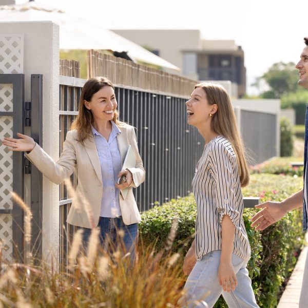 Friendly realtor agent welcoming young excited Hispanic married couple at entrance of modern residential property, showcasing new home for sale or rent. Real estate consultation, homeownership, dream